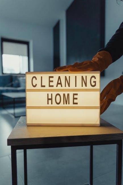 A person wearing yellow rubber gloves is adjusting or setting up a lightbox sign that reads 'CLEANING HOME' in large, bold letters. The sign is illuminated from within, providing a warm glow against a