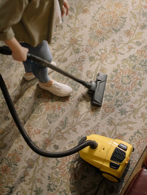 A person operating a yellow vacuum cleaner with a black hose attachment, performing surface cleaning on a traditional patterned carpet in a residential setting. The individual is wearing light-colored