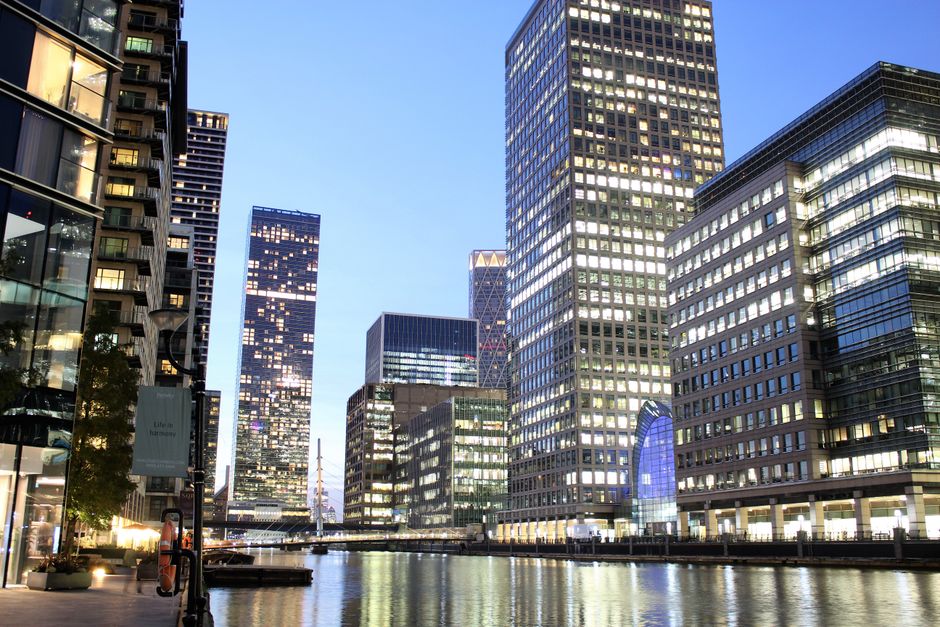 Photograph of a modern office district along a waterway in Canary Wharf, London, displaying tall glass and steel buildings with illuminated windows in the evening sky. The scene features a mix of high