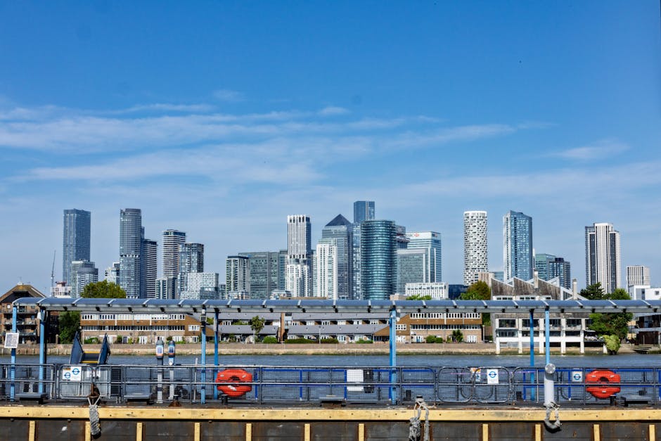 Photograph of a modern office district along a waterway in Canary Wharf, London, displaying tall glass and steel buildings with illuminated windows in the evening sky. The scene features a mix of high